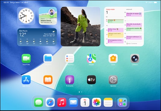 Três modelos de iPad na horizontal com o modelo do meio na frente dos outros dois, Apple Pencil magneticamente fixado à parte superior do modelo do meio; a tela do aparelho do meio mostrando a Tela de Início com ícones de app coloridos com o efeito do Liquid Glass; a tela à esquerda mostrando o app Pré-Visualização e a tela à direita mostrando a Tela de Início com arquivos saindo do Dock em formato de leque