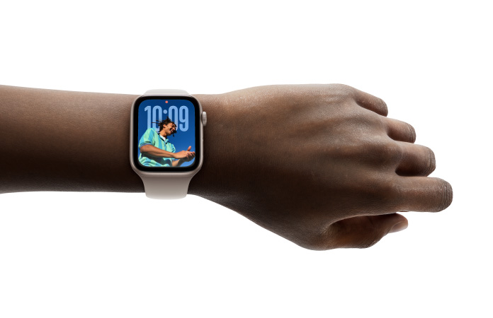 Close do Apple Watch com caixa prateada e pulseira de borracha no pulso de uma pessoa, mostrando o app Hora no novo design com Liquid Glass.