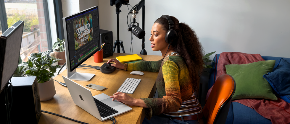Moteris namuose dirba prie stalo naudodama „MacBook Pro“, kuris prijungtas prie dviejų išorinių ekranų, dėvi ausines ir kalba į mikrofoną