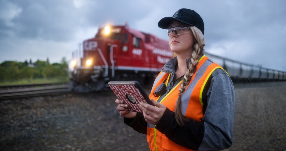Een spoorwegmedewerker houdt een iPad vast en kijkt uit over het spoor terwijl er een trein voorbij rijdt