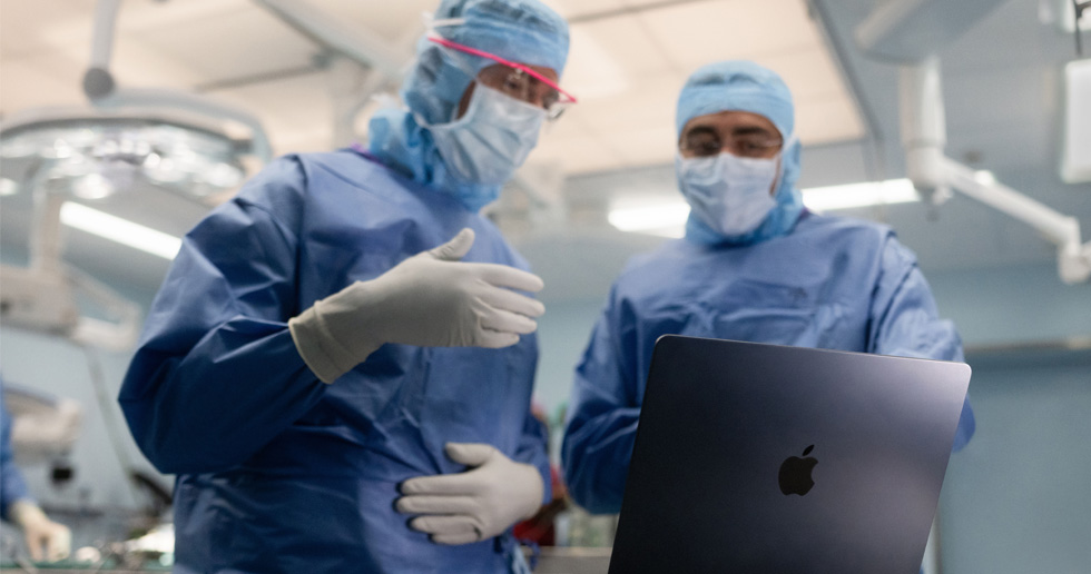 Twee chirurgen in de operatiekamer kijken naar een MacBook, de één gebaart met zijn hand terwijl de ander observeert