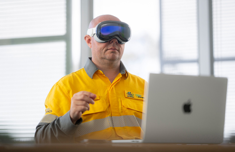 Een medewerker draagt Apple Vision Pro en maakt een knijpend gebaar, terwijl een MacBook Pro in de kleur Silver open op het bureau staat