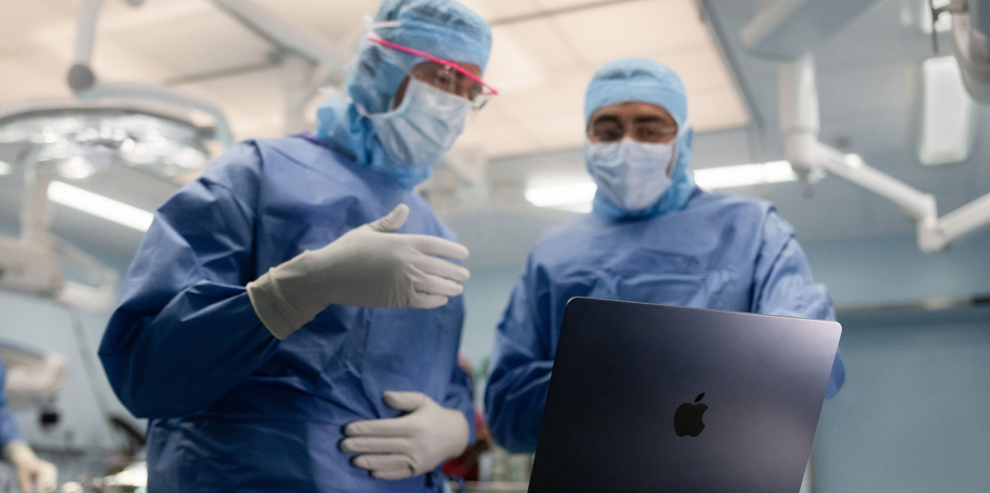 Deux chirurgiens sont dans une salle d’opération et regardent un MacBook; l’un fait un geste de la main tandis que l’autre observe