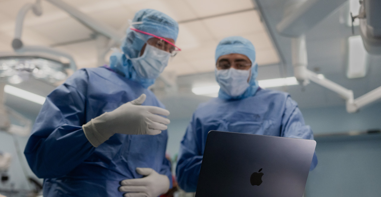 兩位外科醫生在手術室，討論 MacBook Pro 螢幕上的內容。